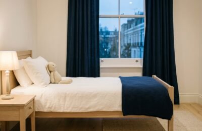 A calm empty child's bedroom at dusk with a bedside lamp lit, a knitted toy rabbit against the pillow and a navy blanket across the bed
